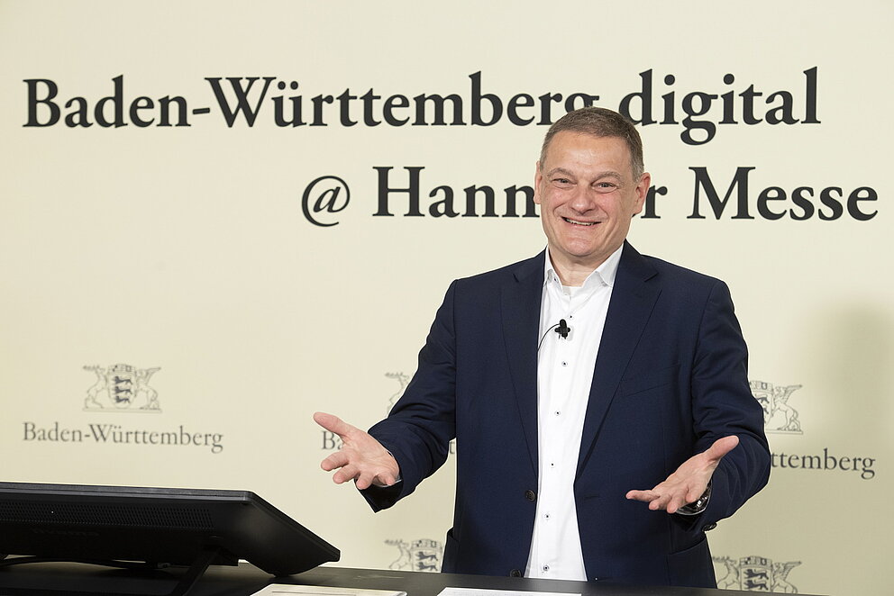 Franz Loogen blickt lächelnd in die Kamera. Er steht vor einer Wand mit dem Schriftzug 'Baden-Württemberg digital @ Hannover Messe'. Seine Hände sind leicht nach vorne ausgestreckt, um die Gäste zu begrüßen.. 