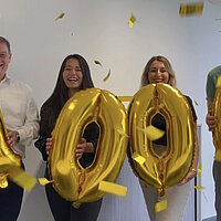 Vier Personen halten goldene Luftballons in die Kamera, die die Zahl 1000 ergeben und im Hintergrund fliegt goldenes Konfetti.