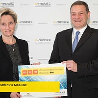 Wirtschaftsministerin Dr. Nicole Hoffmeister-Kraut über gibt Franz Loogen ein Schild, auf dem Mittelstandsoffensive Mobilität zu lesen ist. Beide blicken dabei lächelnd in die Kamera.
