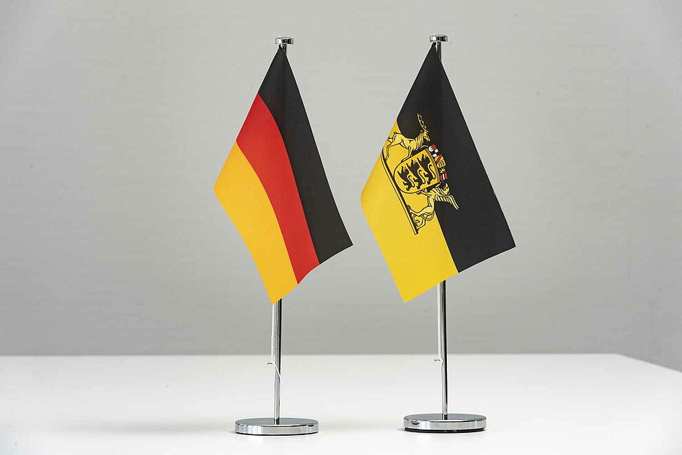 Flaggen von Deutschland und Baden-Württemberg stehen als Wimpel auf einem weißen Tisch.