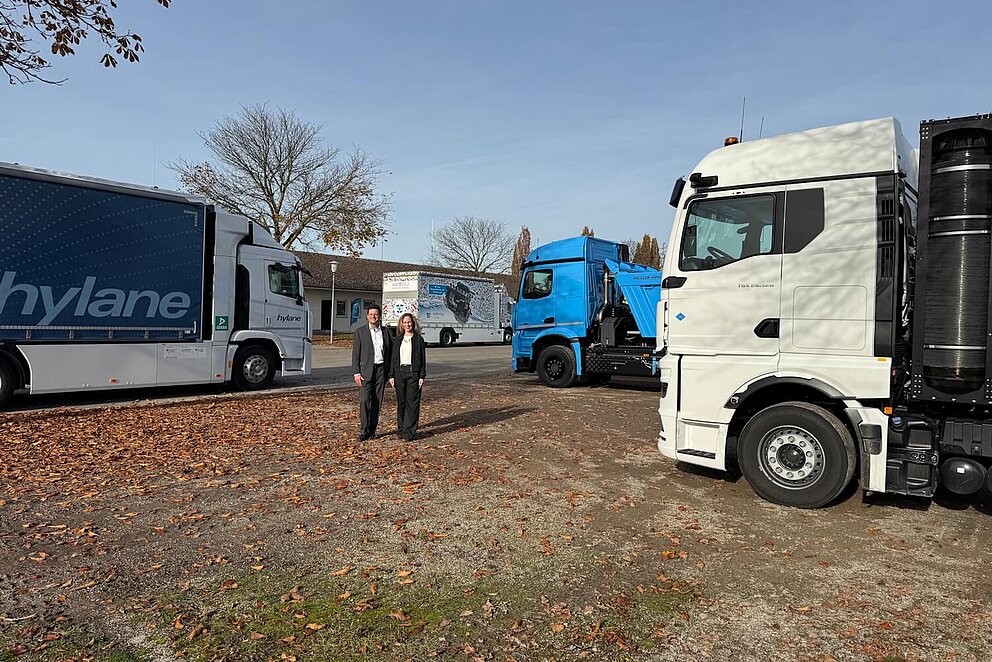 Außenaufnahme von zwei Personen, die in die Kamera lächeln. Im Hintergrund sind Nutzfahrzeuge zu sehen.