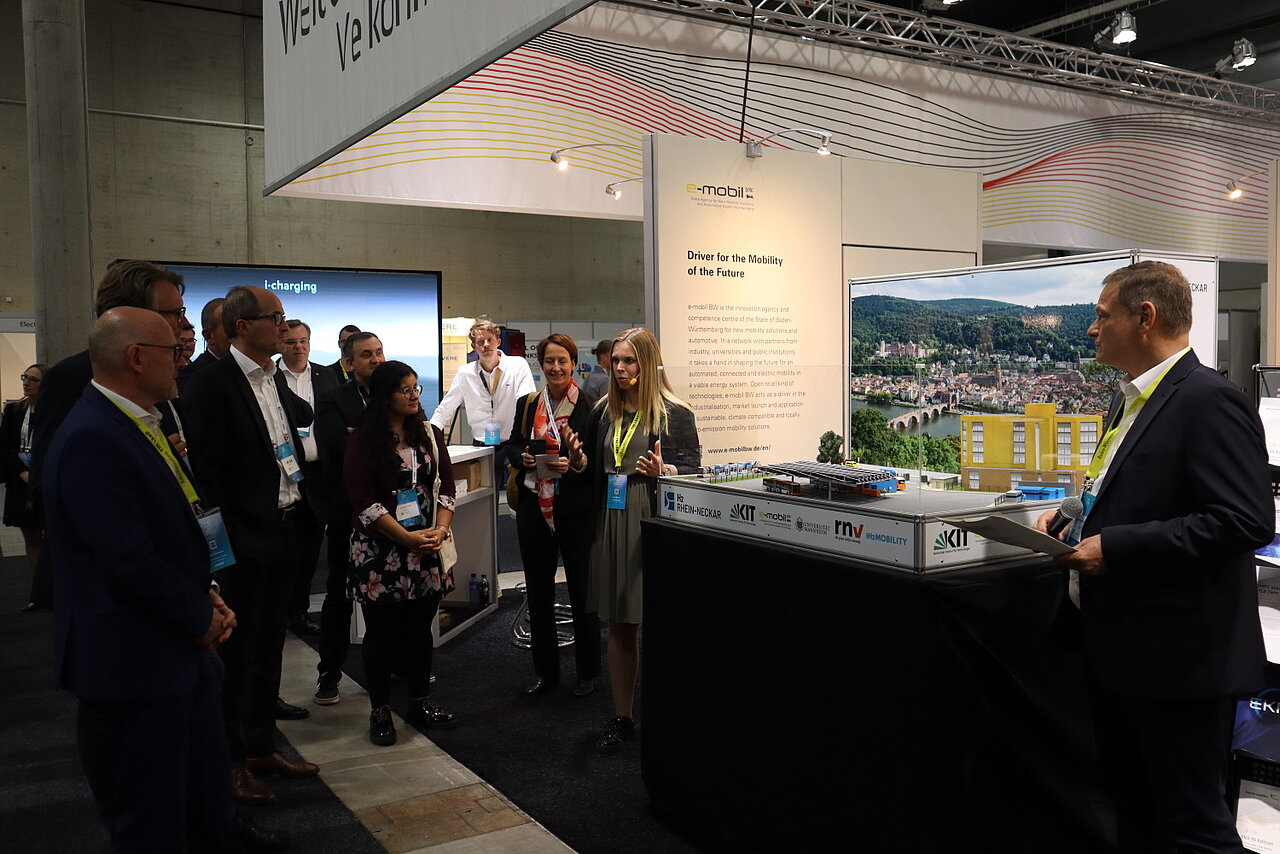 Alina Richter und Franz Loogen haben ein Mikrophon und sprechen zu Verkehrsminister Winfried Hermann MdL beim Messerundgang über den deutschen Gemeinschaftsstand auf der EVS35 in Oslo.