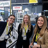 Drei Menschen lächeln in die Kamera. Im Hintergrund sieht man eine Messehalle von Innen.