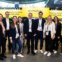 Eine Gruppe Menschen steht auf dem Messestand und lächelt in die Kamera.