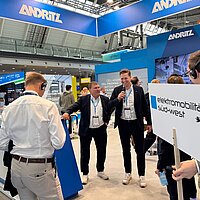 Eine Personengruppe steht auf dem Messestand von Andritz Schuler Pressen und hört zwei Personen zu, die einen Vortrag halten.