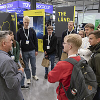 Eine Gruppe Besuchern steht im Kreis zusammen auf einem Messestand.
