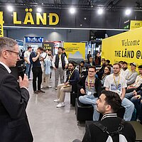 Ein Mann hält eine Rede auf einer Messe. Junge Menschen sitzen auf Sofas vor ihm und hören zu.