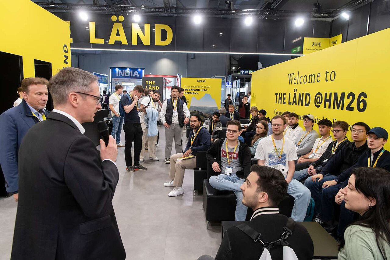 Ein Mann hält eine Rede auf einer Messe. Junge Menschen sitzen auf Sofas vor ihm und hören zu.