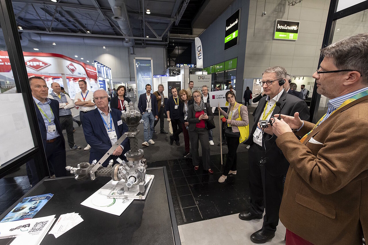 Mehrere Personen stehen um ein Exponat auf der Messe herum, ein Mann erklärt was zu sehen ist.