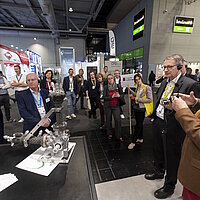 Mehrere Personen stehen um ein Exponat auf der Messe herum, ein Mann erklärt was zu sehen ist.