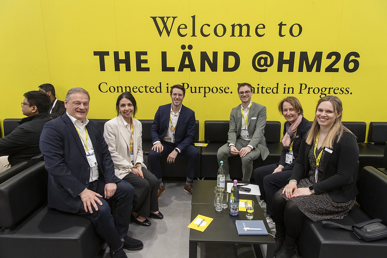 6 Personen sitzen auf schwarzen Sofas vor einer gelben Messewand mit der Aufschrift "Welcome to THE LÄND HM26" und lächeln in die Kamera