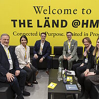 6 Personen sitzen auf schwarzen Sofas vor einer gelben Messewand mit der Aufschrift "Welcome to THE LÄND HM26" und lächeln in die Kamera
