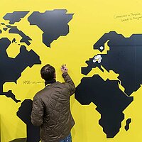 Ein Mann steht vor einer großen Wand mit dem Aufdruck einer Weltkarte und zeichnet etwas ein. 