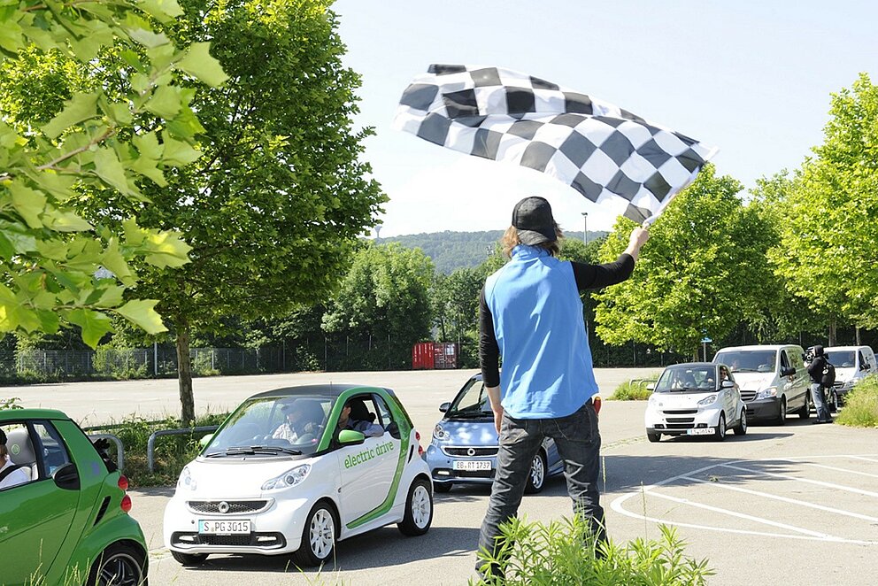 Ein Mann schwingt auf einem Parkplatz eine schwarz-weiß karierte Flagge als Startsignal. Im Hintergrund stehen mehrere Autos in einer Reihe, überwiegend Smart-Modelle. Eines der Autos trägt die Aufschrift ‚electric drive‘.