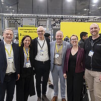 Sechs Personen stehen auf einem Messestand im gelben THE LÄND Design.