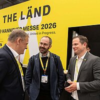 Drei Männer unterhalten sich auf einer Messe. Im Hintergrund sieht man die Aufschrift 'THE LÄND'.