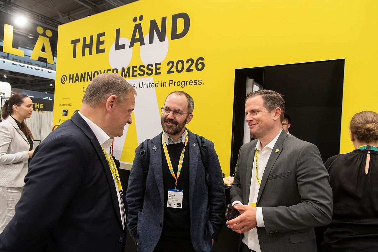 Drei Männer unterhalten sich auf einer Messe. Im Hintergrund sieht man die Aufschrift 'THE LÄND'.