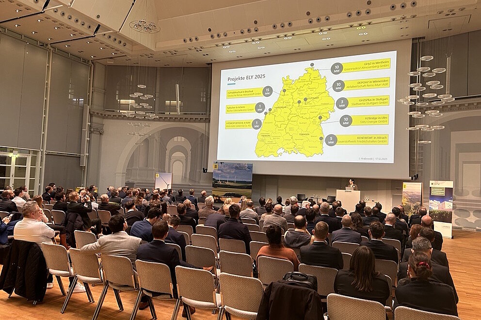 Vortrag mit gelber Folie. Viele Menschen sitzen auf Stühlen davor und hören sich den Vortrag an.