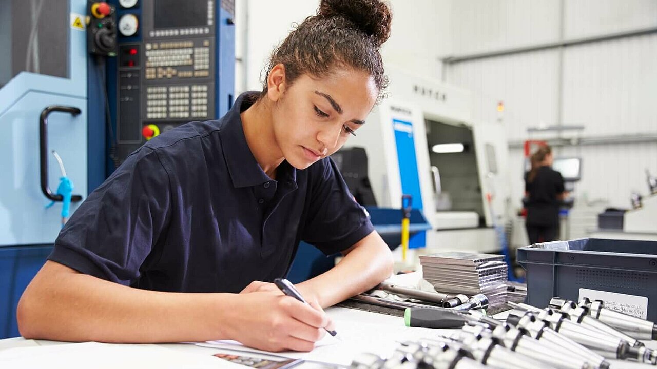 Eine Ingenieurin schreibt, im Hintergrund ist eine CNC-Maschine zu sehen.