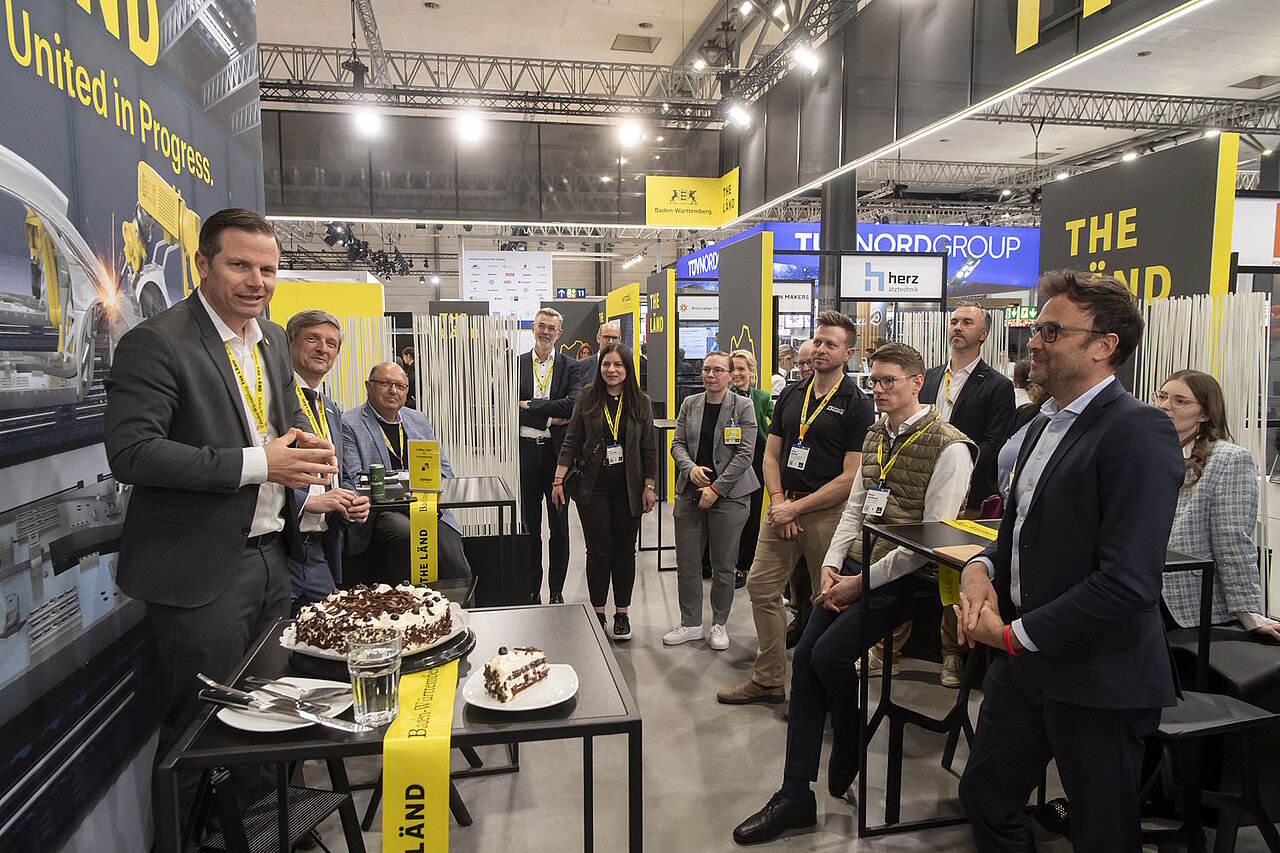 An einem Messestand wird Kuchen serviert, mehrere Personen stehen um den Kuchen herum und hören einem Redner zu.