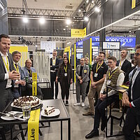 An einem Messestand wird Kuchen serviert, mehrere Personen stehen um den Kuchen herum und hören einem Redner zu.