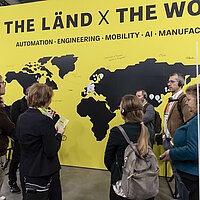 Mehrere Personen stehen vor einer gelben Weltkarte mit der Aufschrift THE LÄND x THE WORLD.