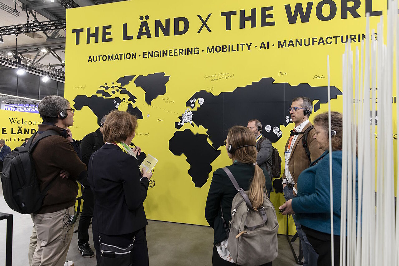 Mehrere Personen stehen vor einer gelben Weltkarte mit der Aufschrift THE LÄND x THE WORLD.