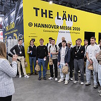 Eine Gruppe junger Erwachsene steht im Halbkreis vor einer gelben Wand mit der Aufschrift "THE LÄND" und hört einer Frau in hellgrauem Blazer zu.