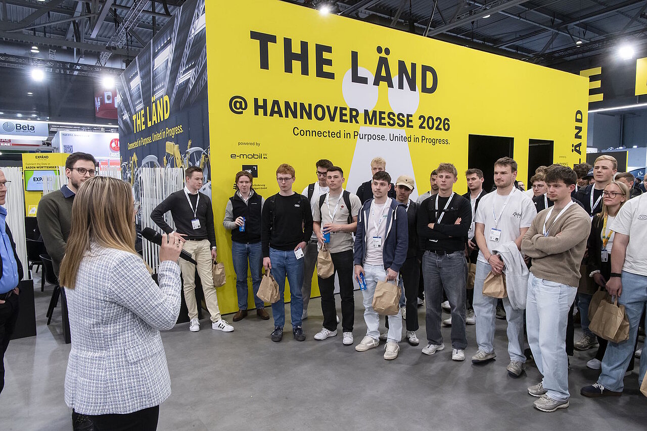 Eine Gruppe junger Erwachsene steht im Halbkreis vor einer gelben Wand mit der Aufschrift "THE LÄND" und hört einer Frau in hellgrauem Blazer zu.