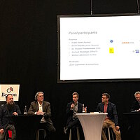 Blick auf die Bühne während einer Paneldiskussion mit einem stehenden Moderator und fünf sitzenden Teilnehmenden aus Finnland und Deutschland. 