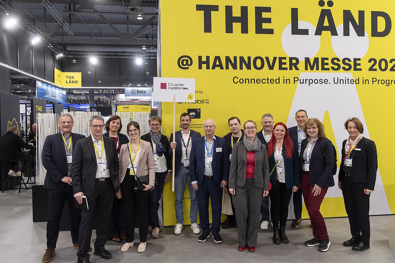 Eine Gruppe Menschen steht vor einer gelben Messewand und hält ein Schild hoch. 