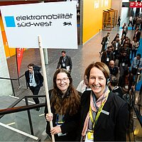 Zwei Frauen halten ein Schild mit dem Titel "Elektromobilität Süd-West".