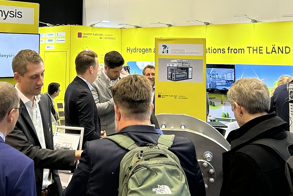 Vier Männer stehen um ein Elektrolyse-Exponat und sprechen miteinander. Im Hintergrund gelbe Wände mit "THE LÄND" Logo.