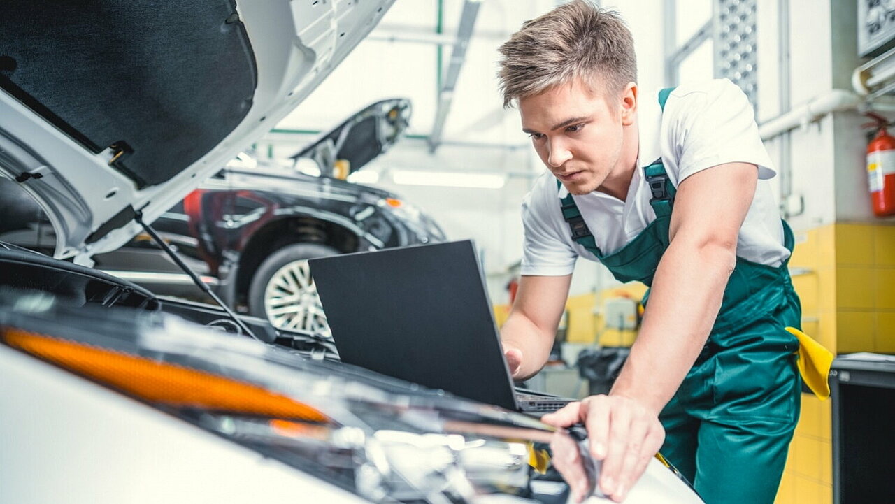 Junger Automechaniker blickt auf Laptop vor einem geöffneten Motorraum.