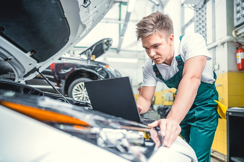 Junger Automechaniker blickt auf Laptop vor einem geöffneten Motorraum.