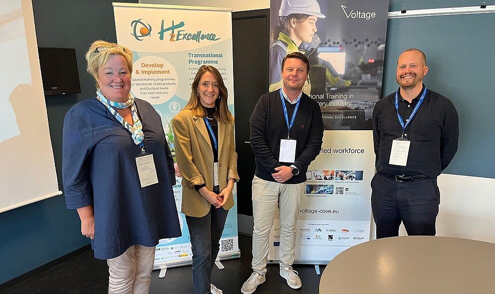 Vier Personen stehen in einem Raum während des Workshops im Rahmen des Forum on Vocational Excellence in Kolding, Dänemark. Links ein Banner des Projekts H2Excellence und rechts ein Banner des Projekts Voltage.