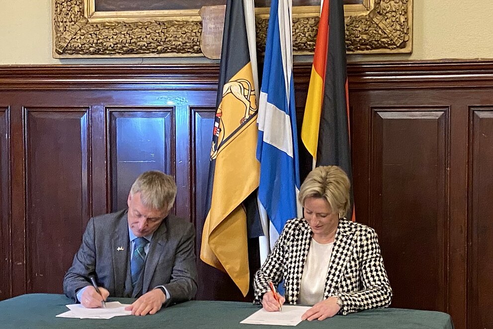 Die beiden Wirtschaftsminister:innen von Baden-Württemberg und Schottland sitzen an einem Tisch und unterzeichnen ein Papier, im Hintergrund sind die Fahnen von Deutschland, Baden-Württemberg und Schottland zu sehen.
