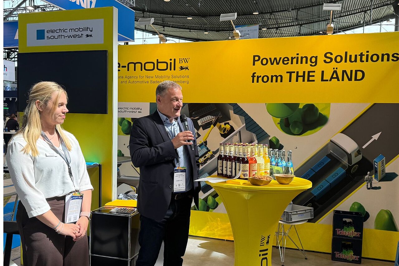 Ein Mann und eine Frau stehen vor einer gelben Messewand der e-mobil BW und der Mann spricht in ein Mikrofon.