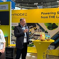Ein Mann und eine Frau stehen vor einer gelben Messewand der e-mobil BW und der Mann spricht in ein Mikrofon.