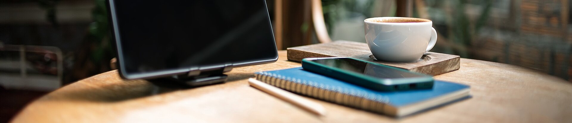 Ein Tablet steht neben einem blauen Notizblock, einem Handy und einer Tasse Kaffee auf einem runden Holztisch. Im Hintergrund sieht man einen Holzstuhl, eine Pflanze und eine Fensterfront.