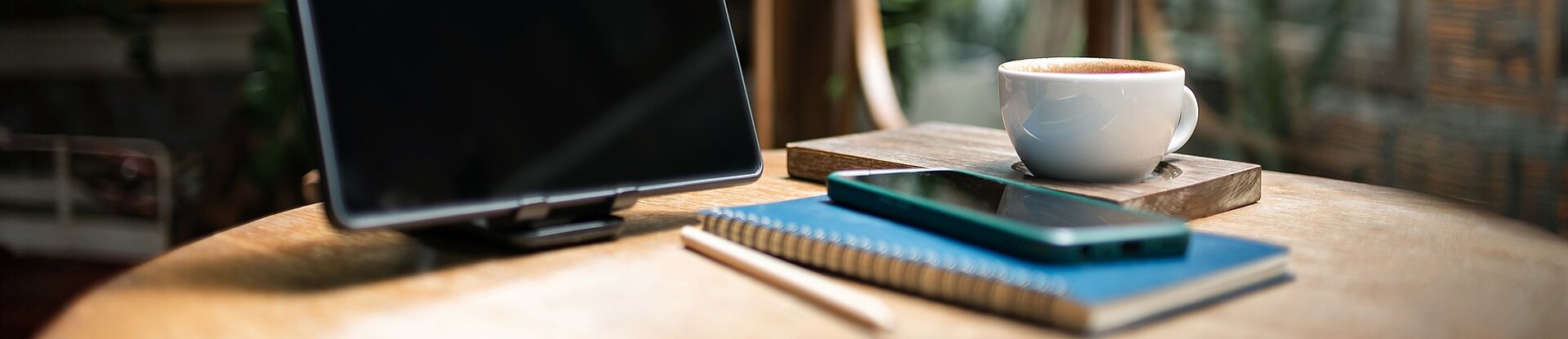 Ein Tablet steht neben einem blauen Notizblock, einem Handy und einer Tasse Kaffee auf einem runden Holztisch. Im Hintergrund sieht man einen Holzstuhl, eine Pflanze und eine Fensterfront.