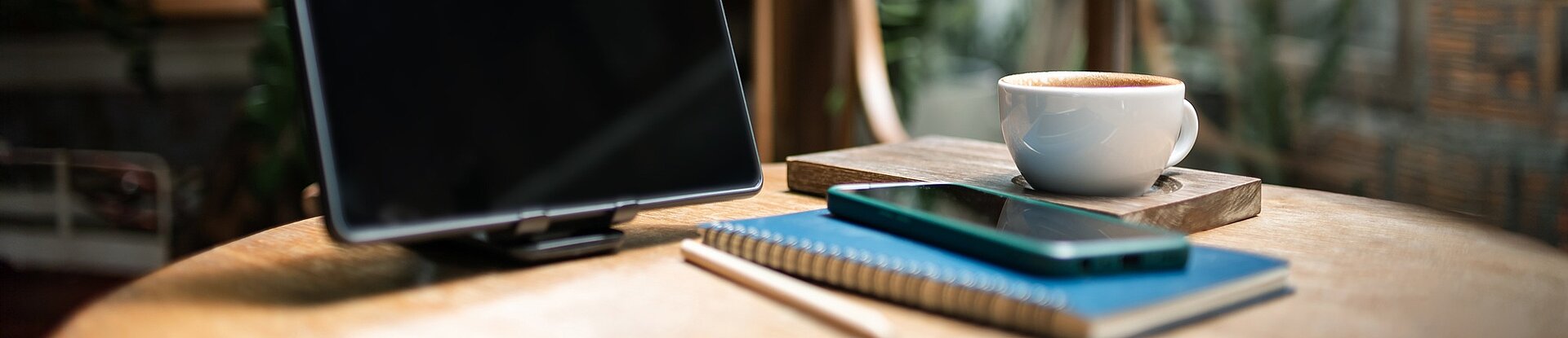 Tablet, Notizblock und Handy in einem Cafe Ein Tablet steht neben einem blauen Notizblock, einem Handy und einer Tasse Kaffee auf einem runden Holztisch. Im Hintergrund sieht man einen Holzstuhl, eine Pflanze und eine Fensterfront.