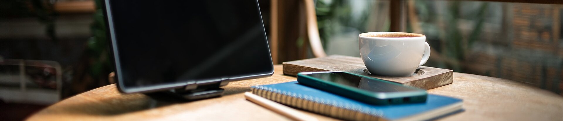 Ein Tablet steht neben einem blauen Notizblock, einem Handy und einer Tasse Kaffee auf einem runden Holztisch. Im Hintergrund sieht man einen Holzstuhl, eine Pflanze und eine Fensterfront.
