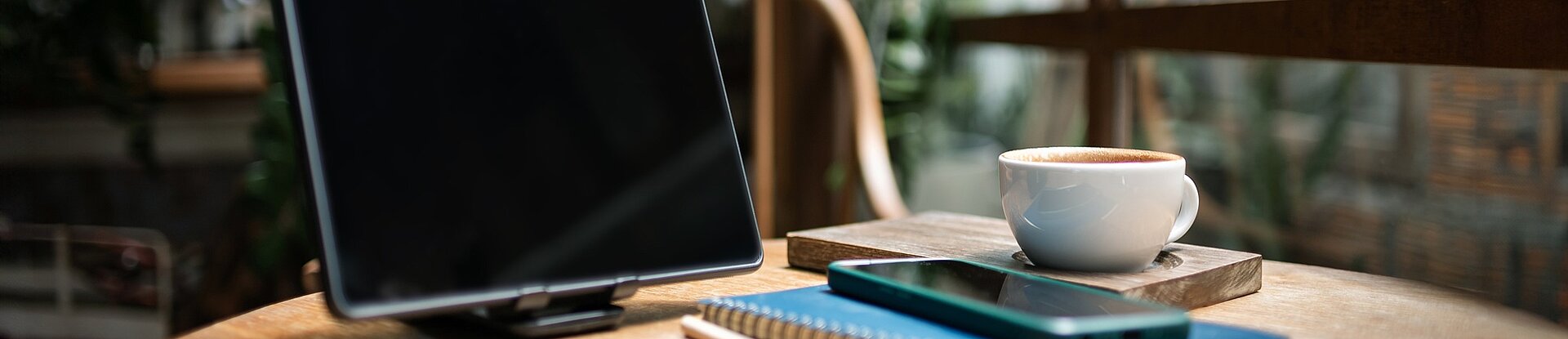 Ein Tablet steht neben einem blauen Notizblock, einem Handy und einer Tasse Kaffee auf einem runden Holztisch. Im Hintergrund sieht man einen Holzstuhl, eine Pflanze und eine Fensterfront.