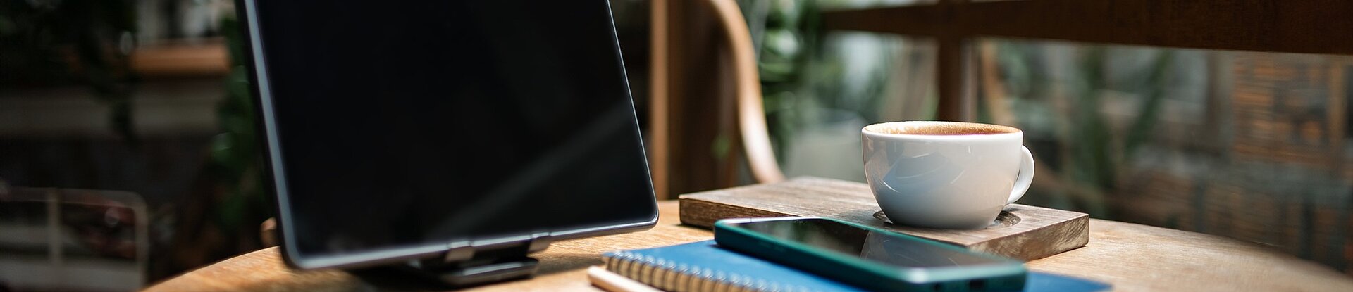 Ein Tablet steht neben einem blauen Notizblock, einem Handy und einer Tasse Kaffee auf einem runden Holztisch. Im Hintergrund sieht man einen Holzstuhl, eine Pflanze und eine Fensterfront.