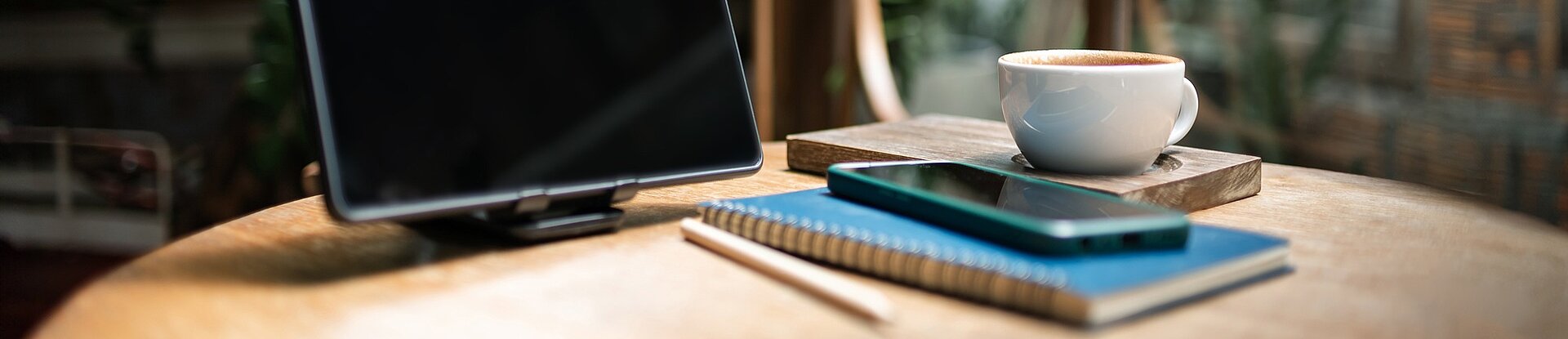 Tablet, Notizblock und Handy in einem Cafe Ein Tablet steht neben einem blauen Notizblock, einem Handy und einer Tasse Kaffee auf einem runden Holztisch. Im Hintergrund sieht man einen Holzstuhl, eine Pflanze und eine Fensterfront.