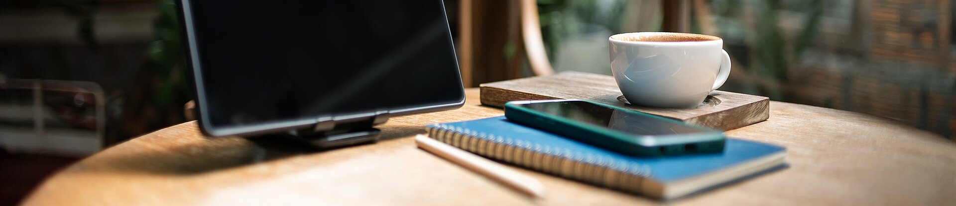 Tablet, Notizblock und Handy in einem Cafe Ein Tablet steht neben einem blauen Notizblock, einem Handy und einer Tasse Kaffee auf einem runden Holztisch. Im Hintergrund sieht man einen Holzstuhl, eine Pflanze und eine Fensterfront.