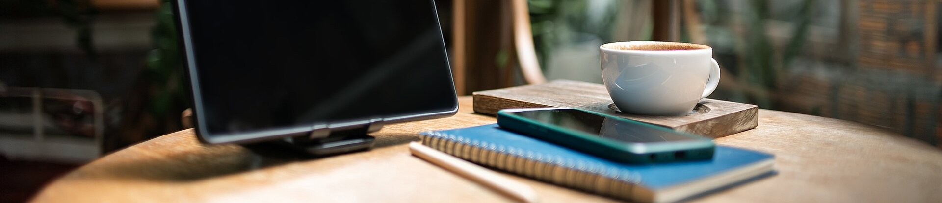 Ein Tablet steht neben einem blauen Notizblock, einem Handy und einer Tasse Kaffee auf einem runden Holztisch. Im Hintergrund sieht man einen Holzstuhl, eine Pflanze und eine Fensterfront.