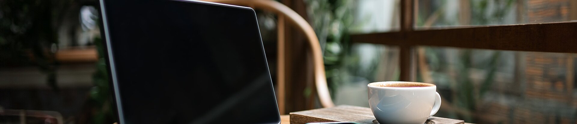 Ein Tablet steht neben einem blauen Notizblock, einem Handy und einer Tasse Kaffee auf einem runden Holztisch. Im Hintergrund sieht man einen Holzstuhl, eine Pflanze und eine Fensterfront.