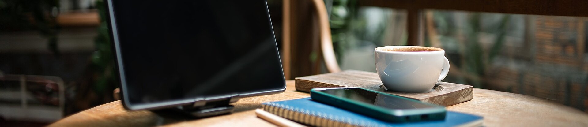 Ein Tablet steht neben einem blauen Notizblock, einem Handy und einer Tasse Kaffee auf einem runden Holztisch. Im Hintergrund sieht man einen Holzstuhl, eine Pflanze und eine Fensterfront.